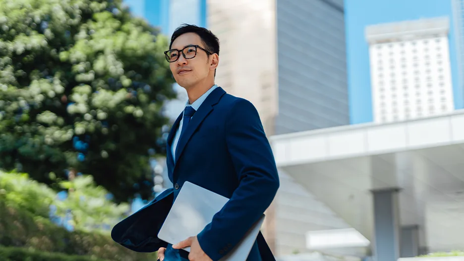 A man wearing a suit, walking outside his office carrying a laptop