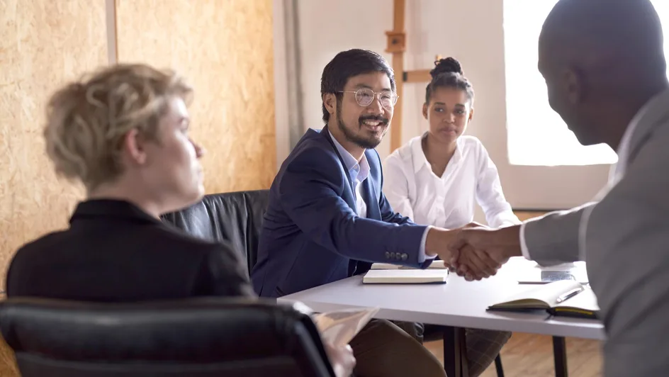 Smiling Asian businessman shaking hands with an African colleague during a meeting at a table in an office board room