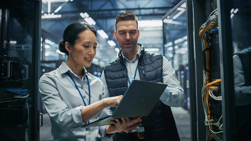 A man and a woman discussing something while looking at a laptop screen in a factory