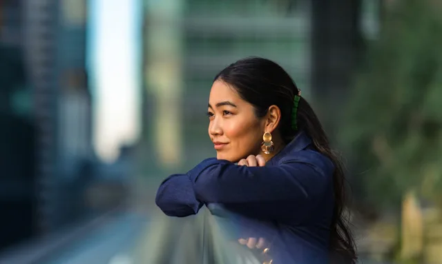 A woman leans against a railing, gazing at the city skyline in the background.