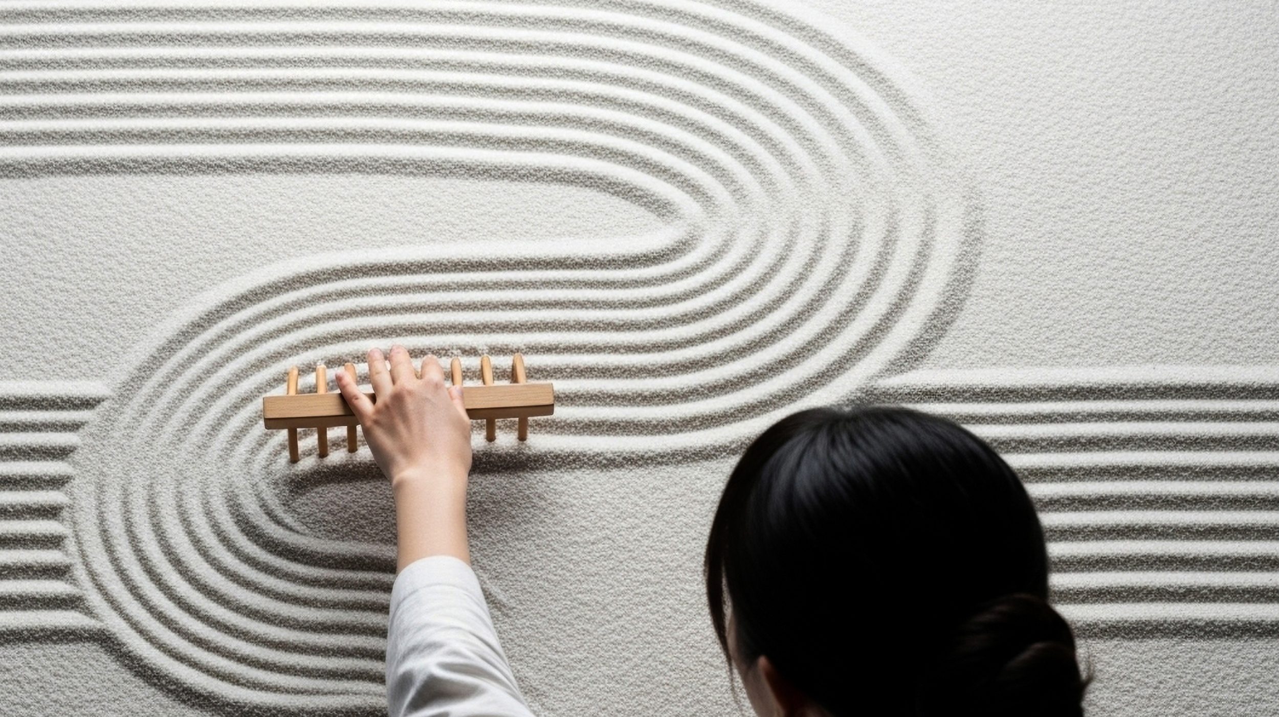 A female figure creating abstract curved lines creating a wave-like pattern in textured sand
