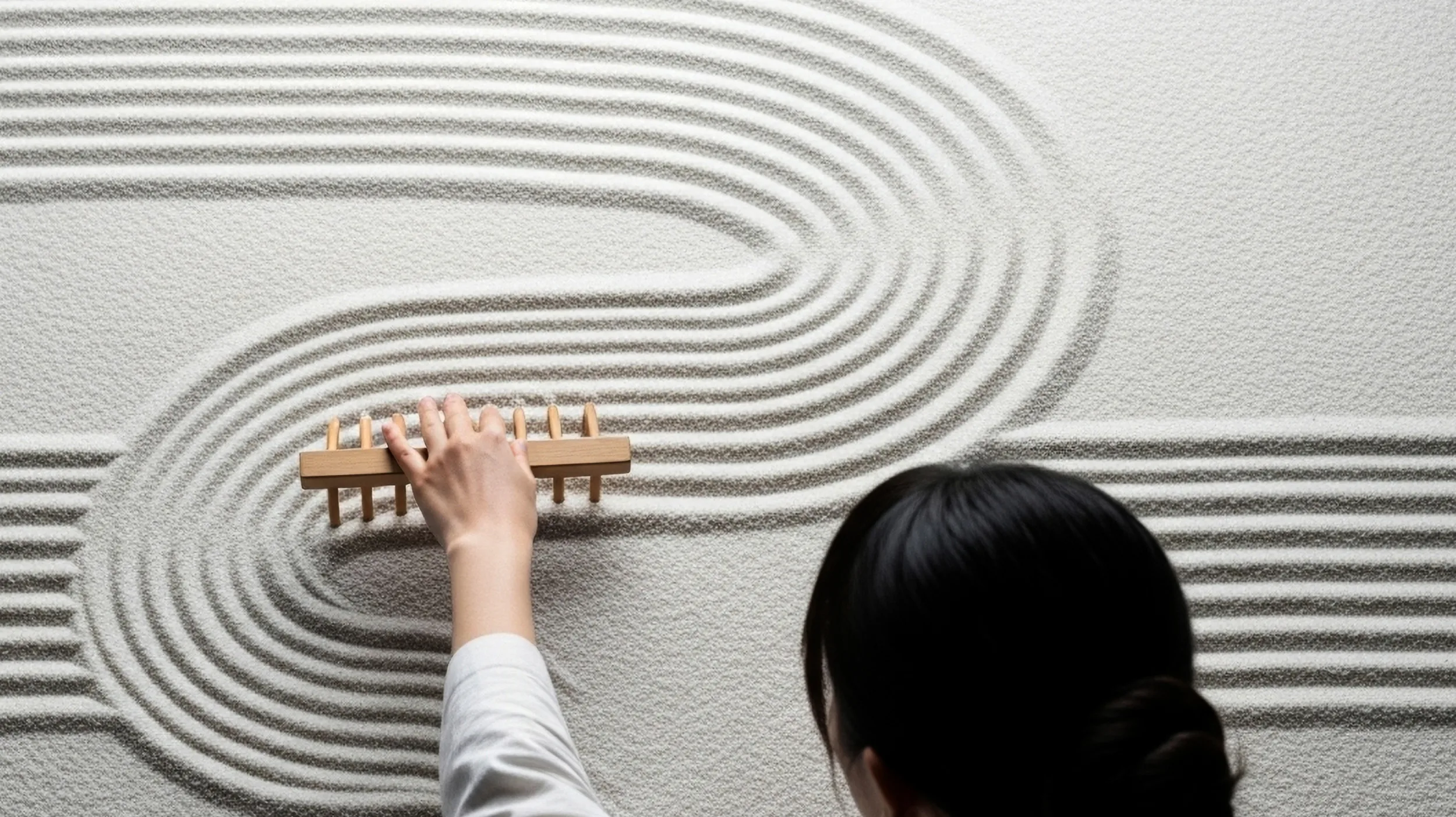 A female figure creating abstract curved lines creating a wave-like pattern in textured sand