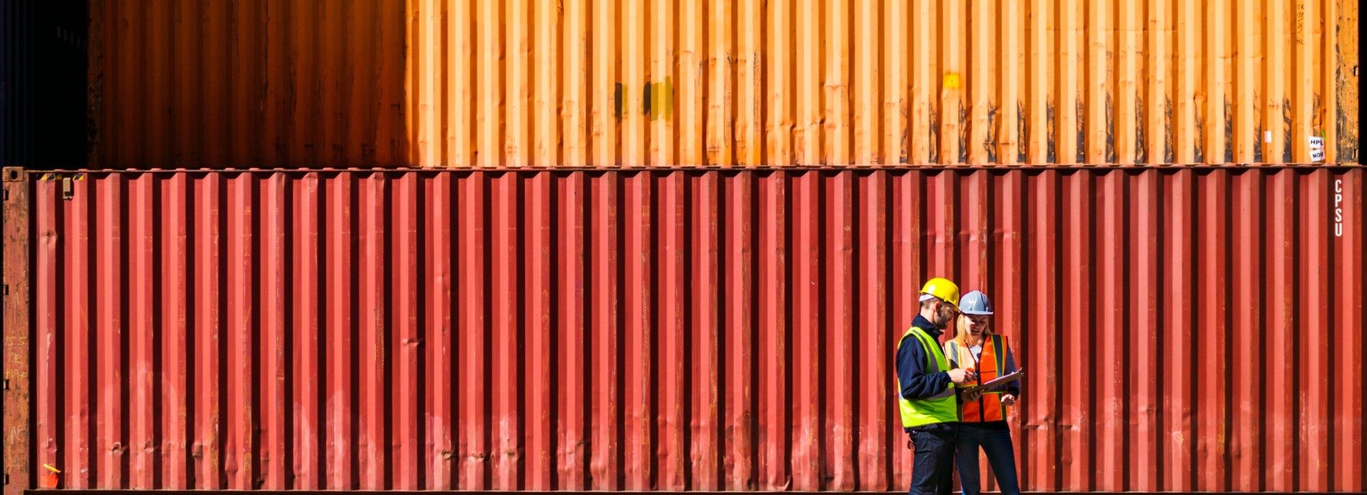 two people in hard hats talking in front of a stack of shipping containers