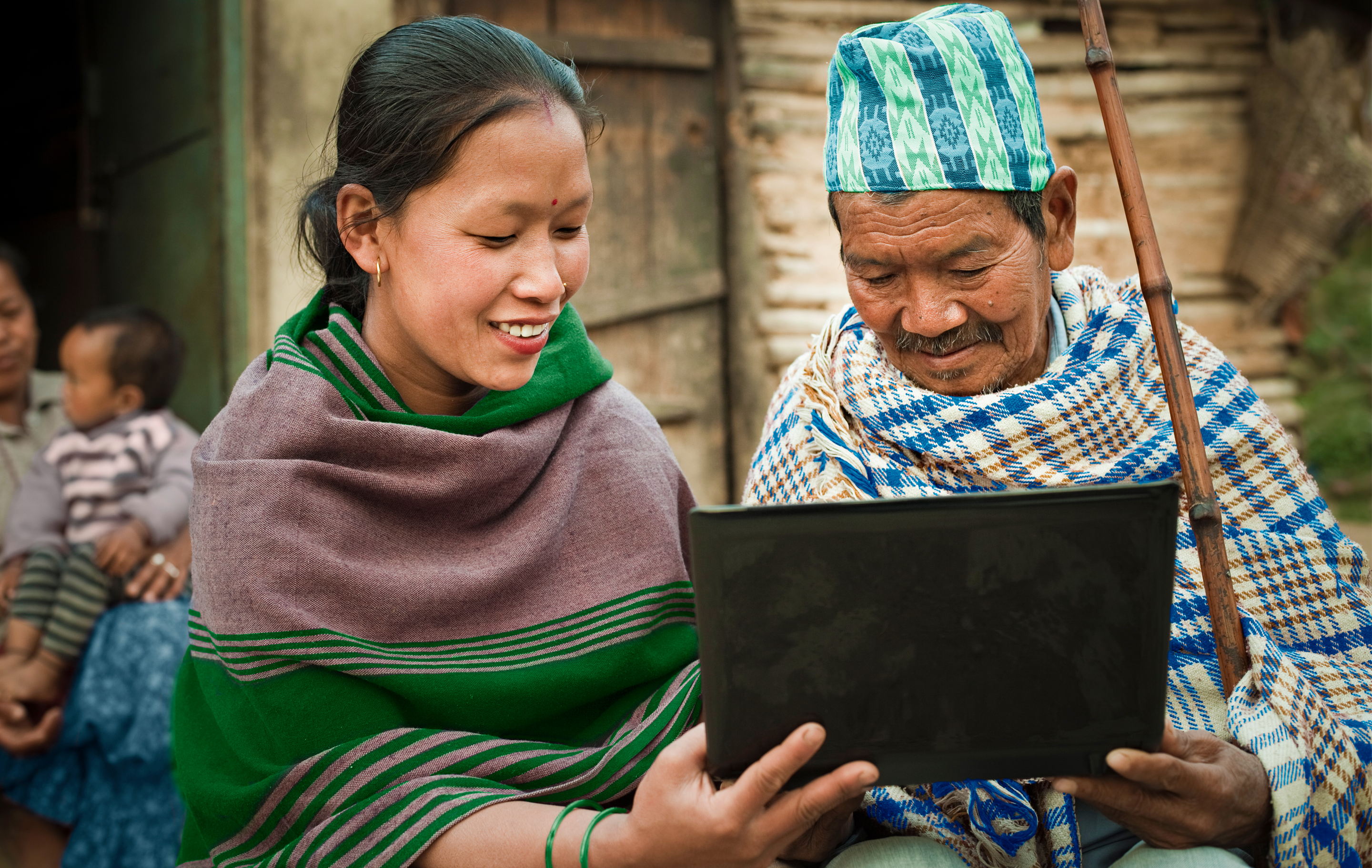 A woman helps a man use a laptop. They're in a rural village and both wearing traditional dress.