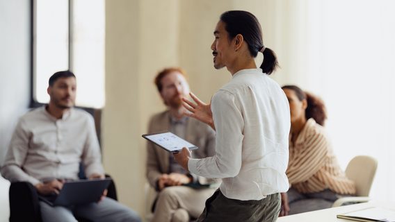 A diverse group of people engaged in a team meeting, with a presenter using a digital tablet in an office environment.