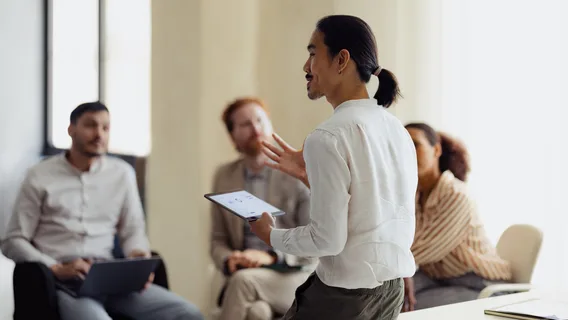 A diverse group of people engaged in a team meeting, with a presenter using a digital tablet in an office environment.